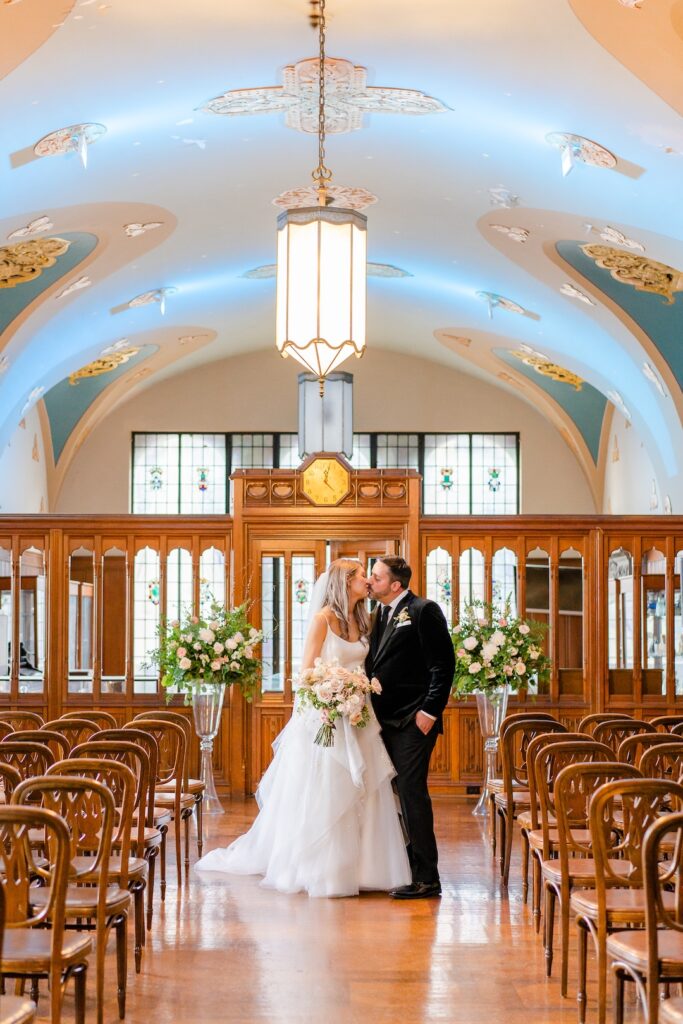 Bride and groom kiss in elegant wedding venue with floral decor and arched ceilings.