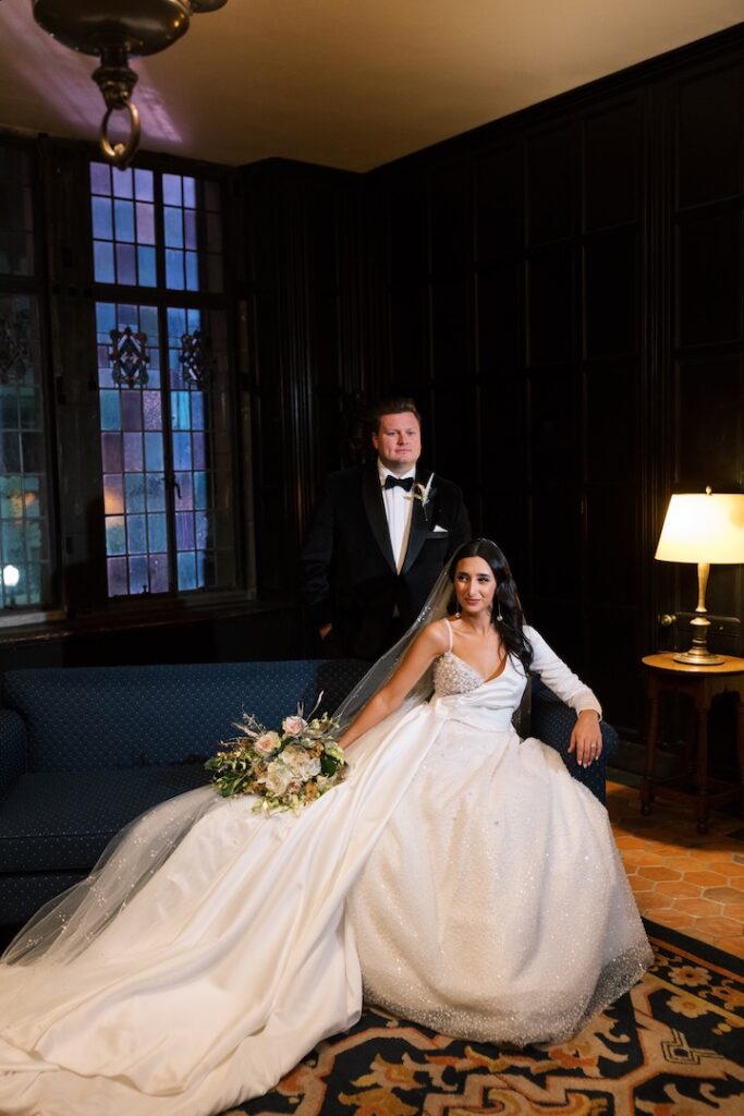 Bride in elegant gown with groom in tuxedo, seated on blue sofa in dimly lit room with bouquet.
