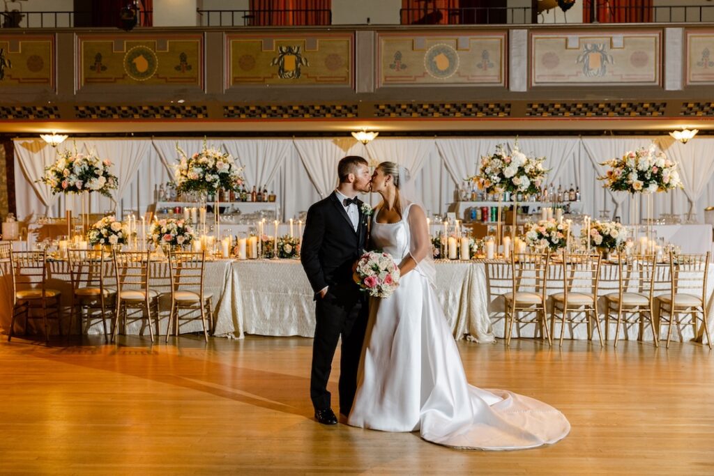 Bride and groom kissing in elegant wedding venue with floral decor and candlelit reception table.