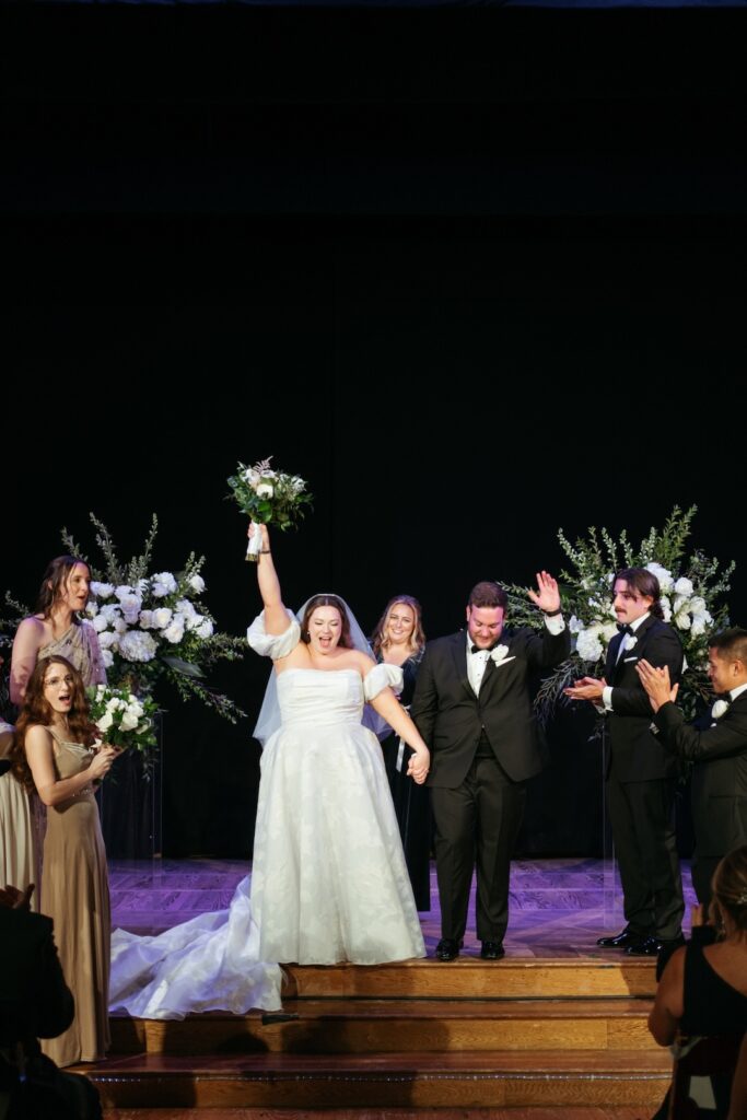 Bride and groom celebrate on stage with joyous wedding party, surrounded by vibrant floral decor.