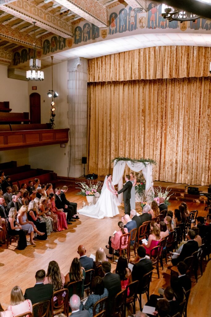 Elegant indoor wedding ceremony with couple exchanging vows, surrounded by guests in formal attire.