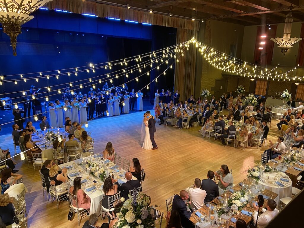 Elegant wedding ballroom dance under string lights, surrounded by guests and a live band.