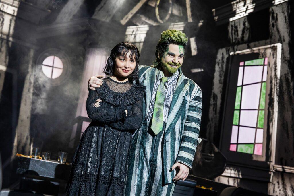 Two actors in theatrical costumes on stage, one in a green striped suit and the other in a black dress.