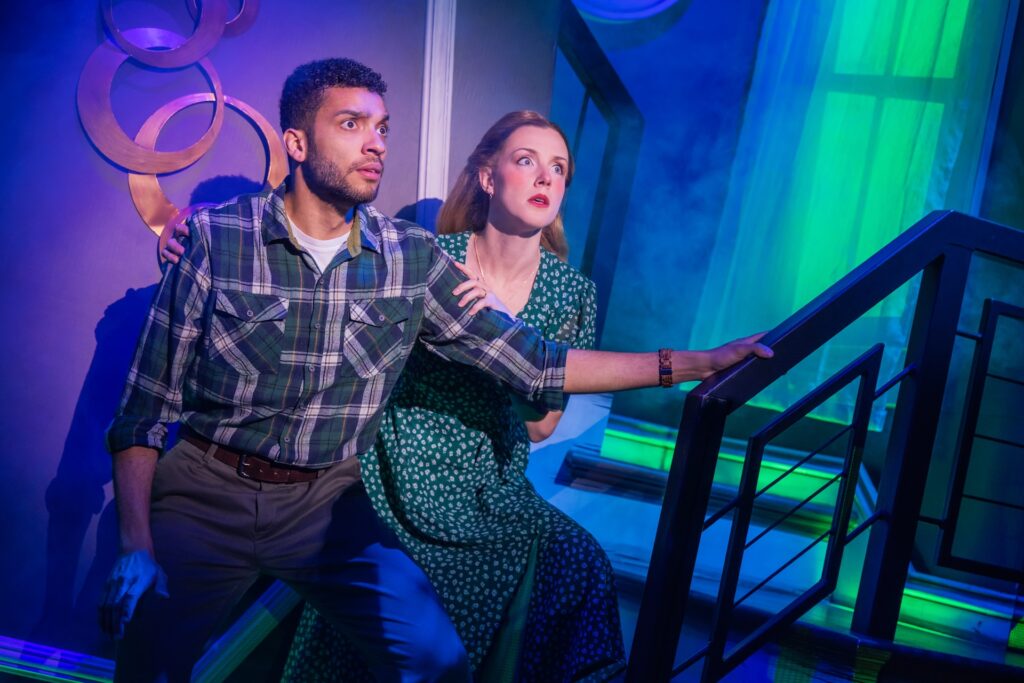 Actors on a dimly lit stage, looking surprised and holding onto a staircase railing.