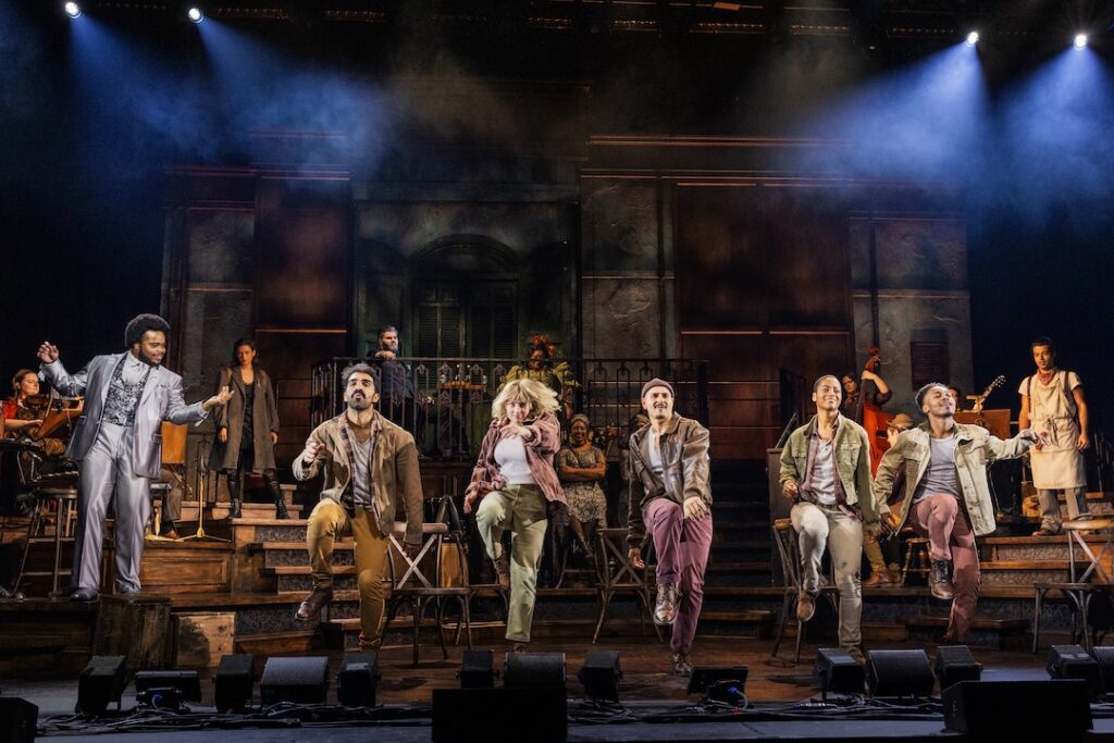 Cast performing a lively musical number on stage with dramatic lighting and set design.