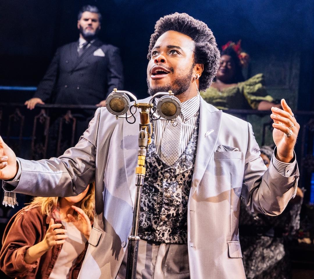 Performer in a suit sings passionately with a vintage microphone on stage, supported by cast members in the background.