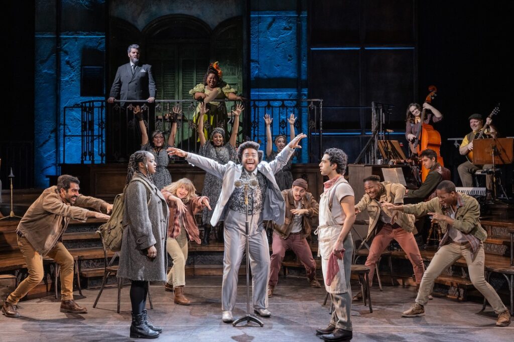 Stage performance with a diverse cast singing and dancing, featuring musicians in the background.