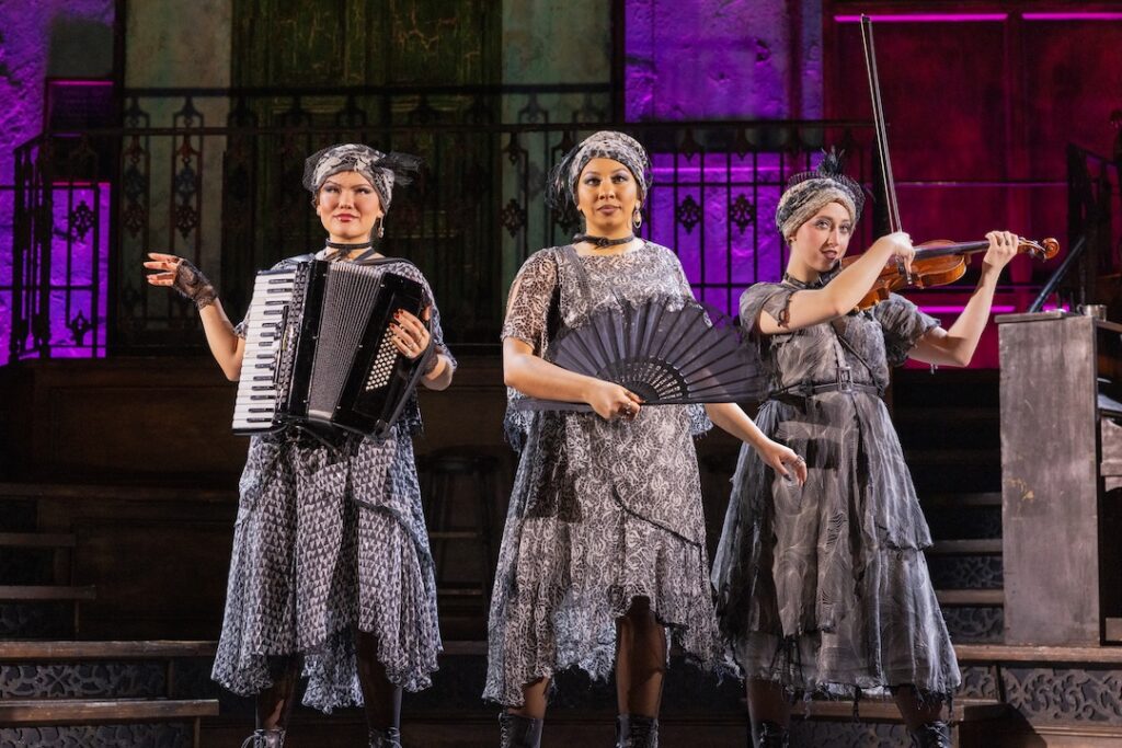 Three performers in vintage outfits playing accordion, holding a fan, and a violin on a theatrical stage.