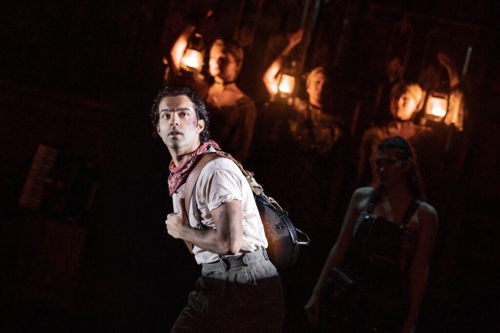 Theatrical performance with actor holding guitar, dramatic lighting, and background figures holding lanterns.