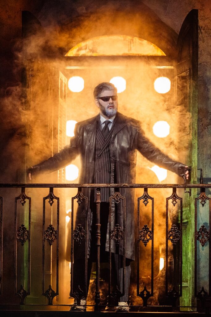 Man in a leather coat stands confidently on a smoky balcony, backlit by bright lights.