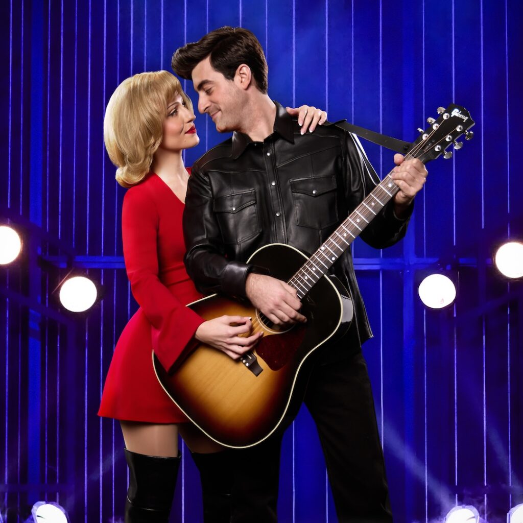 Couple in vintage attire embracing with guitar on stage, romantic atmosphere with blue and purple lights.